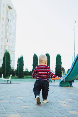 Sarışın bir çocuk, 1 yaşında, çocuk parkında yürüyor. Çocuk temiz havada oyun oynuyor. Çocuk gelişimi. Kot ve spor ayakkabılı bir çocuk..