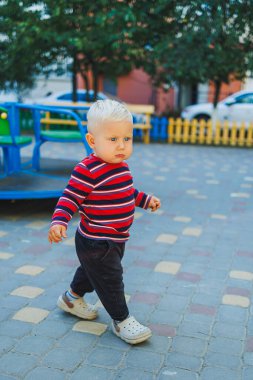 Sarışın bir çocuk, 1 yaşında, çocuk parkında yürüyor. Çocuk temiz havada oyun oynuyor. Çocuk gelişimi. Kot ve spor ayakkabılı bir çocuk..