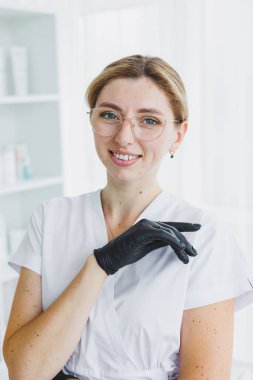 Gözlüklü ve lateks eldivenli dermatolog. İyi ışıklandırılmış ofisi olan beyaz önlüklü profesyonel bir kozmetik uzmanı..