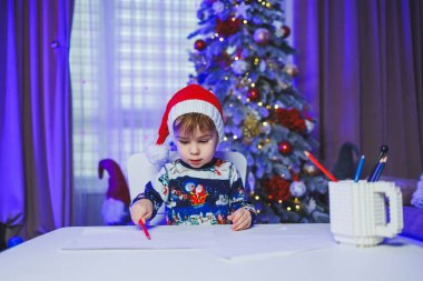 Küçük bir çocuk evde, masada oturuyor ve bir Noel ağacının arka planında oynuyor. Evde Noel ağacı var. Bir çocuğun yeni yıl havası..