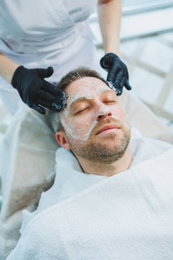 Bir güzellik uzmanı bir adamın yüzüne tıbbi maske takar. Erkekler için yüz bakımı. Erkek kozmetolojisi.