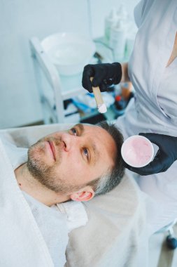 Güzellik salonunda maskeli bir adam. Erkeklerin yüz bakımı konsepti. Güzellik uzmanı bir kaplıca merkezinde müşterinin yüzüne maske takar. Erkek kozmetolojisi.