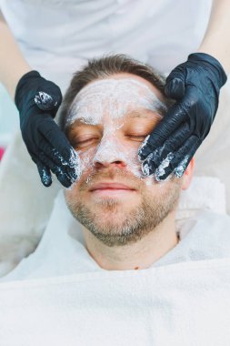 Bir güzellik uzmanı bir adamın yüzüne tıbbi maske takar. Erkekler için yüz bakımı. Erkek kozmetolojisi.