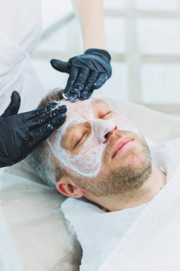 Güzellik salonunda maskeli bir adam. Erkeklerin yüz bakımı konsepti. Güzellik uzmanı bir kaplıca merkezinde müşterinin yüzüne maske takar. Erkek kozmetolojisi.