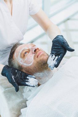 Güzellik salonunda maskeli bir adam. Erkeklerin yüz bakımı konsepti. Güzellik uzmanı bir kaplıca merkezinde müşterinin yüzüne maske takar. Erkek kozmetolojisi.