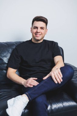 Siyah tişörtlü genç yakışıklı bir adam siyah bir kanepede oturuyor. Bir adam rahat bir kanepede dinlenir..