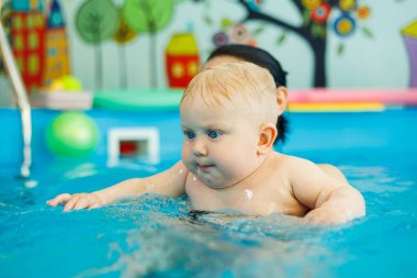 Bir bebek havuzda koçla yüzmeyi öğrenir. Erken yaşta havuzda yüzmek. Kapalı havuzda oynayan ve eğlenen kadın eğitmeni olan mutlu bir çocuk..
