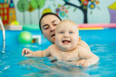 Bir bebek havuzda koçla yüzmeyi öğrenir. Erken yaşta havuzda yüzmek. Kapalı havuzda oynayan ve eğlenen kadın eğitmeni olan mutlu bir çocuk..