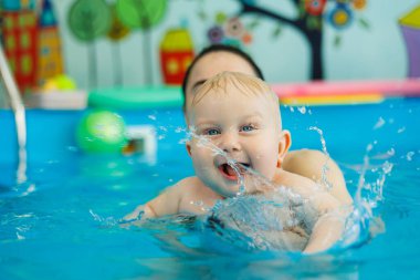 Bir bebek havuzda koçla yüzmeyi öğrenir. Erken yaşta havuzda yüzmek. Kapalı havuzda oynayan ve eğlenen kadın eğitmeni olan mutlu bir çocuk..