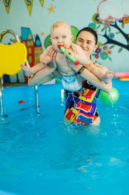 Bir bebek havuzda koçla yüzmeyi öğrenir. Erken yaşta havuzda yüzmek. Kapalı havuzda oynayan ve eğlenen kadın eğitmeni olan mutlu bir çocuk..