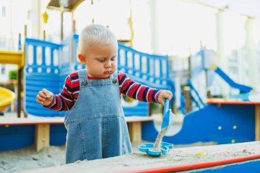 Kot pantolon giymiş 1 yaşındaki bir çocuk kum havuzunda dururken kumla oynuyor. Bir çocuk kum havuzunda otururken plastik raketlerle oynar..