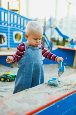 Kot pantolon giymiş 1 yaşındaki bir çocuk kum havuzunda dururken kumla oynuyor. Bir çocuk kum havuzunda otururken plastik raketlerle oynar..