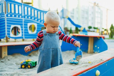 Kot pantolon giymiş 1 yaşındaki bir çocuk kum havuzunda dururken kumla oynuyor. Bir çocuk kum havuzunda otururken plastik raketlerle oynar..