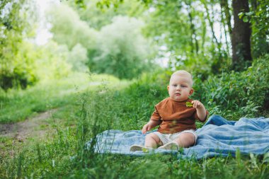 Tişörtlü ve şortlu sarışın bir çocuk yeşil çimlerin üzerinde oturur ve oynar. Sokakta bir çocuk oynuyor. Bebek kendi başına oturuyor..
