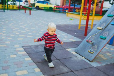 Kotlu ve spor ayakkabılı, sarışın bir çocuk oyun bahçesinde yürüyor. Çocuk temiz havada gelişir ve oynar..
