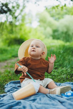 Parktaki çimenlerde oturan küçük bir çocuk şapkalı ve gülümsüyor. Çocuk parkta çimlerin üzerinde oturuyordu. Hasır şapkalı bir çocuk..