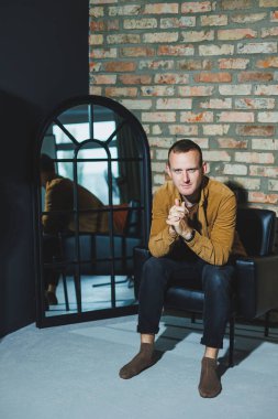 Yakışıklı genç bir adam siyah bir sandalyede oturur ve kameraya bakar. Tuğla duvarın arkasında bir adam.
