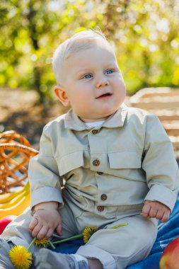 Parktaki çimlerin üzerinde oturan sevimli bir oğlan çocuğu gülümsüyor. Bej tulumlu şık bir bebek..