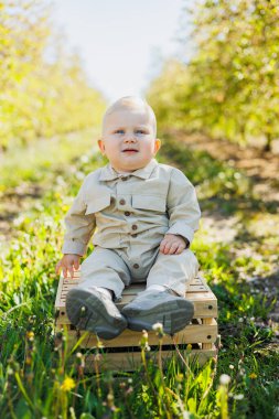 Parktaki çimlerin üzerinde oturan sevimli bir oğlan çocuğu gülümsüyor. Bej tulumlu şık bir bebek..