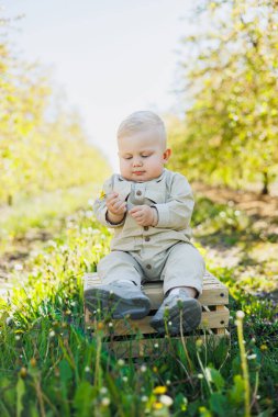 Parktaki çimlerin üzerinde oturan sevimli bir oğlan çocuğu gülümsüyor. Bej tulumlu şık bir bebek..