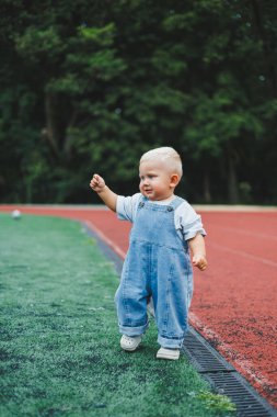 Tişört ve kot pantolon giyen bir yaşında sarışın bir çocuk temiz havada yürür. Çocuğun kişisel gelişimi.