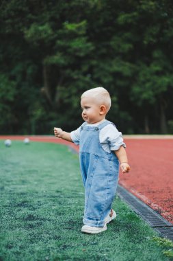 Tişört ve kot pantolon giyen bir yaşında sarışın bir çocuk temiz havada yürür. Çocuğun kişisel gelişimi.