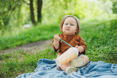 1 yaşında bir çocuk çimlerin üzerinde oturur ve su içer. Yazın temiz havada bir bebek yürüyüşü. Bir çocuk için piknik.