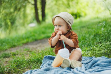 1 yaşında bir çocuk çimlerin üzerinde oturur ve su içer. Yazın temiz havada bir bebek yürüyüşü. Bir çocuk için piknik.