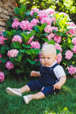 Bir yaşında bir çocuk yeşil çimlerin üzerinde oturuyor. Erkekler için tulumlar.