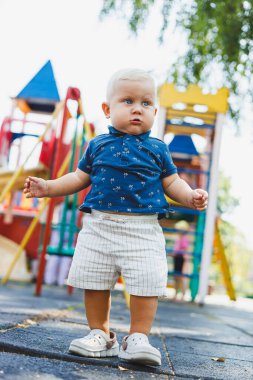 Oyun parkında mavi tişörtlü ve şortlu küçük bir çocuk koşuyor. Çocuk aktif olarak oynayarak ve temiz havada yürüyerek zaman geçirir..