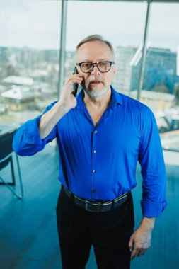 Gömlekli sakallı yaşlı bir adam ofiste çalışıyor ve telefonda konuşuyor. Elinde telefon olan gözlüklü bir işadamı.