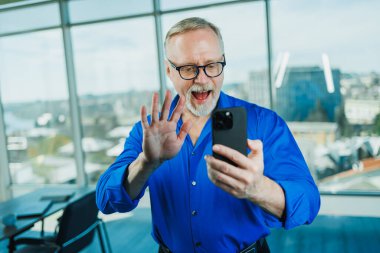 A bearded old man in a shirt works in the office and talks on the phone. A businessman in glasses with a phone in his hands clipart