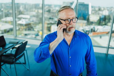 A bearded old man in a shirt works in the office and talks on the phone. A businessman in glasses with a phone in his hands clipart