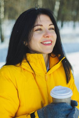 Karlı bir parkın arka planında kahve içen mutlu kadın. Paket kahve. Sıcak kahveyle parkta kış yürüyüşü.