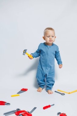Beyaz arka planda pamuk tulumunun içinde plastik oyuncaklarla oynayan 1 yaşında küçük bir çocuk. Bebek oyuncak çekiçle oynuyor..