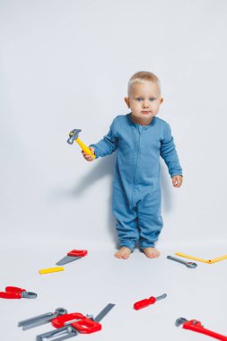 Beyaz arka planda pamuk tulumunun içinde plastik oyuncaklarla oynayan 1 yaşında küçük bir çocuk. Bebek oyuncak çekiçle oynuyor..