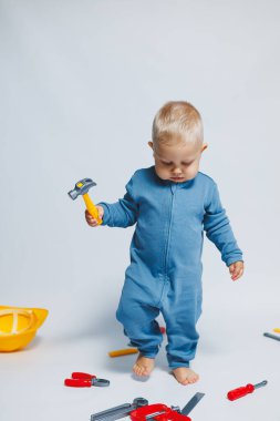 Beyaz arka planda pamuk tulumunun içinde plastik oyuncaklarla oynayan 1 yaşında küçük bir çocuk. Bebek oyuncak çekiçle oynuyor..