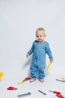 Beyaz arka planda pamuk tulumunun içinde plastik oyuncaklarla oynayan 1 yaşında küçük bir çocuk. Bebek oyuncak çekiçle oynuyor..