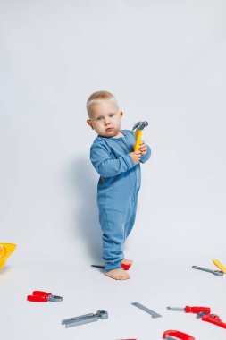 Beyaz arka planda pamuk tulumunun içinde plastik oyuncaklarla oynayan 1 yaşında küçük bir çocuk. Bebek oyuncak çekiçle oynuyor..