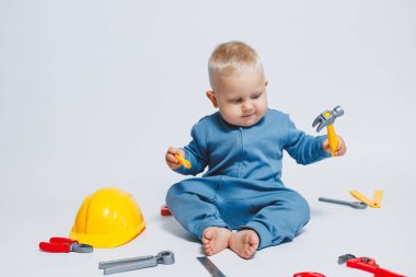 Beyaz arka planda pamuk tulumunun içinde plastik oyuncaklarla oynayan 1 yaşında küçük bir çocuk. Bebek oyuncak çekiçle oynuyor..