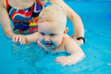 Bir bebek havuzda koçla yüzmeyi öğrenir. Erken yaşta havuzda yüzmek. Kapalı havuzda oynayan ve eğlenen kadın eğitmeni olan mutlu bir çocuk..