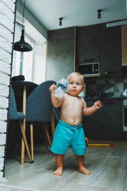 Sarışın bir çocuk evin içinde dolanıyor ve su şişesinden su içiyor. Çocuklar için bir su şişesi..