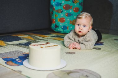 Sarışın bir bebek evde yerde sürünüyor ve tatlı bir pasta yemek istiyor. Çocuk gelişimi. Bir çocuk emeklemeyi öğrenir.