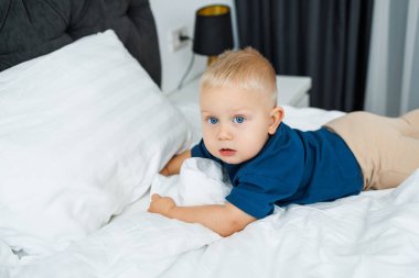 Mavi tişörtlü sarışın bir çocuk beyaz çarşaflı bir yatakta yatıyor. Bebek yatakta otururken oynar. Mavi gözlü küçük bir çocuk.