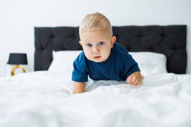 Mavi tişörtlü sarışın bir çocuk beyaz çarşaflı bir yatakta yatıyor. Bebek yatakta otururken oynar. Mavi gözlü küçük bir çocuk.