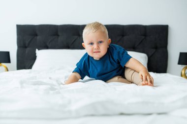 Mavi tişörtlü sarışın bir çocuk beyaz çarşaflı bir yatakta yatıyor. Bebek yatakta otururken oynar. Mavi gözlü küçük bir çocuk.