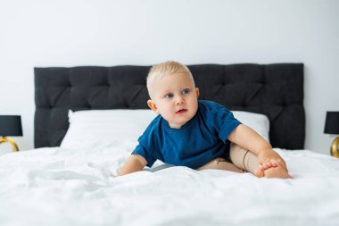 Mavi tişörtlü sarışın bir çocuk beyaz çarşaflı bir yatakta yatıyor. Bebek yatakta otururken oynar. Mavi gözlü küçük bir çocuk.