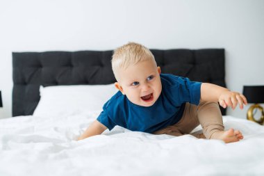 Mavi tişörtlü sarışın bir çocuk beyaz çarşaflı bir yatakta yatıyor. Bebek yatakta otururken oynar. Mavi gözlü küçük bir çocuk.