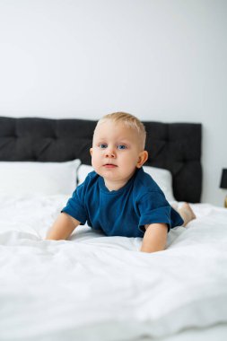 Mavi tişörtlü sarışın bir çocuk beyaz çarşaflı bir yatakta yatıyor. Bebek yatakta otururken oynar. Mavi gözlü küçük bir çocuk.