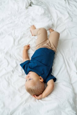 Mavi tişörtlü sarışın bir çocuk beyaz çarşaflı bir yatakta yatıyor. Bebek yatakta otururken oynar. Mavi gözlü küçük bir çocuk.